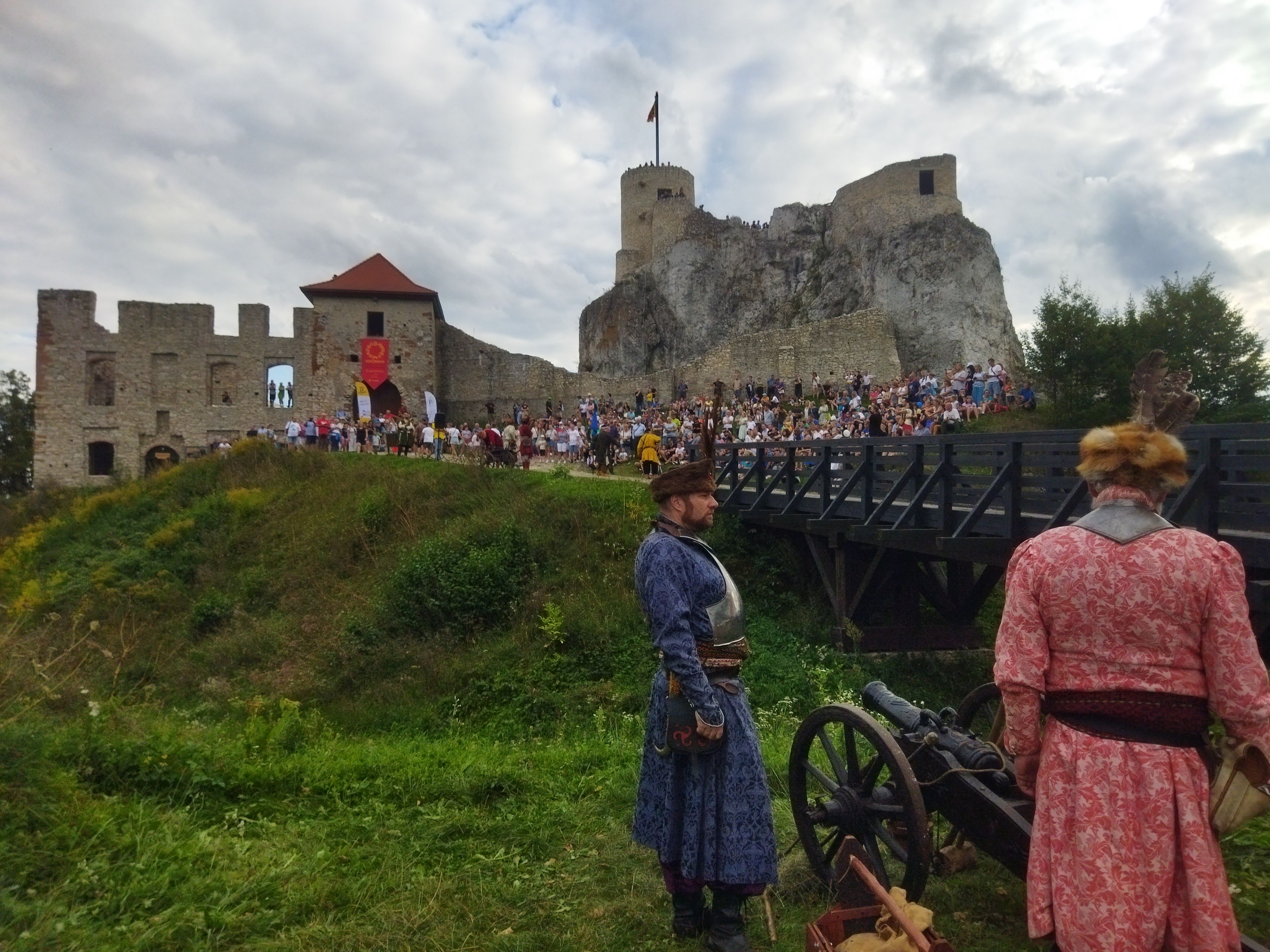 Dwie osoby w dawnych strojach stoją przy armacie. W tle zamek