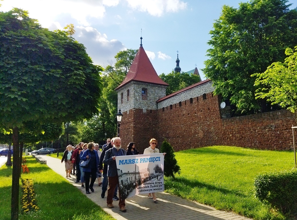 Grupa osób idzie z transparentem z napisem Marsz pamięci. Za nimi czerwona baszta i mury obronne Olkusza.
