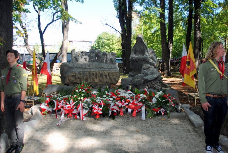 Pomnik Pieta Katyńska na olkuskim cmentarzu. Przed pomnikiem wiązamki kwiatów. Obok pomnika stoi dwóch harcerzy w mundurach.