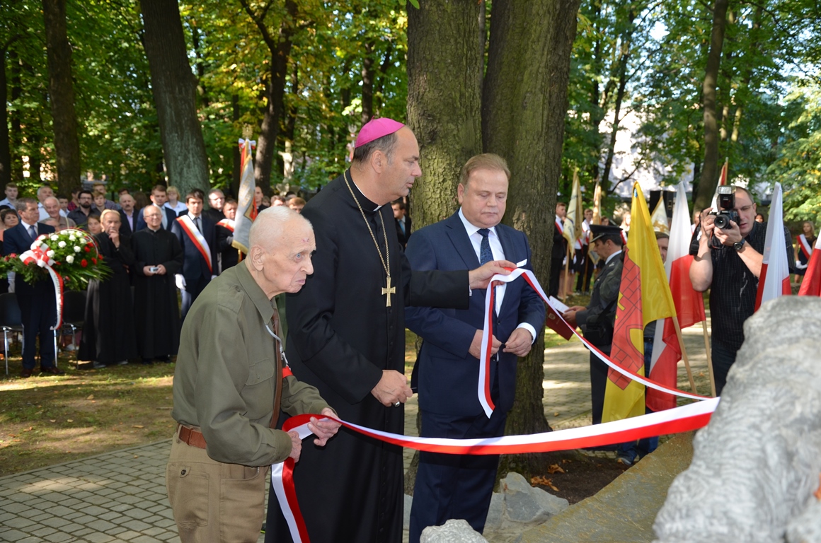 Trzech mężczyzn odsłania pomnik Pieta Katyńska na cmentarzu w Olkuszu. Trzymają w rękach biało czerwone wstęgi. Za nimi grupa osób.
