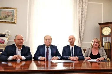 Cztery osoby w eleganckich strojach biznesowych siedzą za drewnianym stołem w reprezentacyjnym pomieszczeniu z zasłonami, zegarem stojącym i obrazem na ścianie.